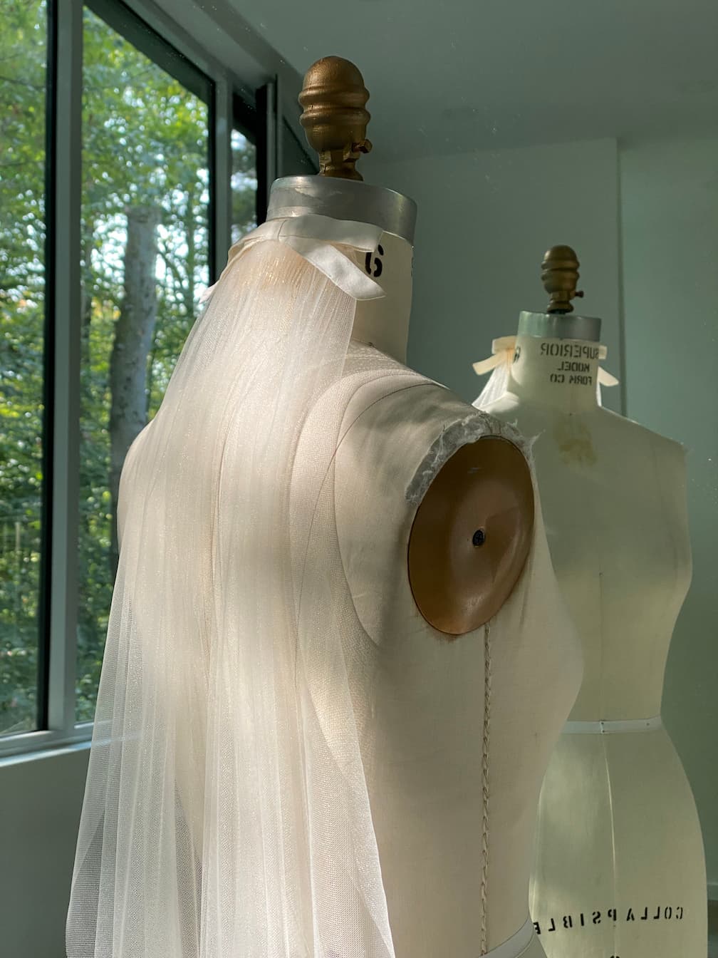 A Noémiah Bow Veil, draped elegantly and topped with a silk bow, stands by a sunlit window. Its sheer white fabric catches the light, reflecting softly in a mirror with a tranquil view of trees outside.