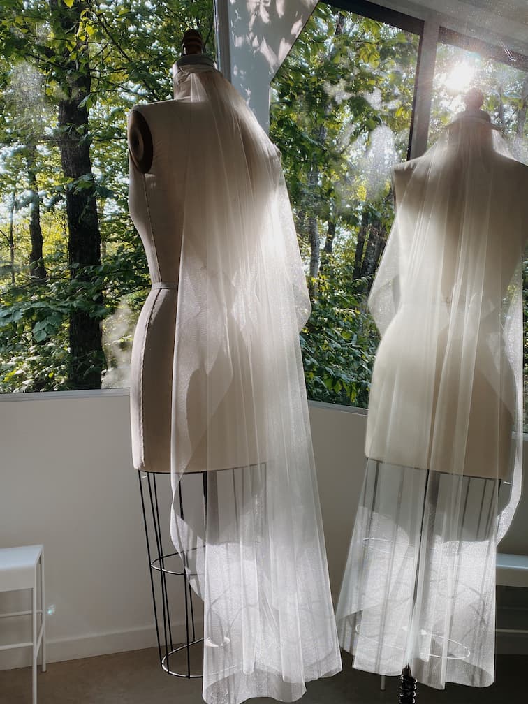 A Noémiah Bow Veil drapes elegantly over a dress form by the window, catching soft sunlight and delicate shadows filtering through the trees.