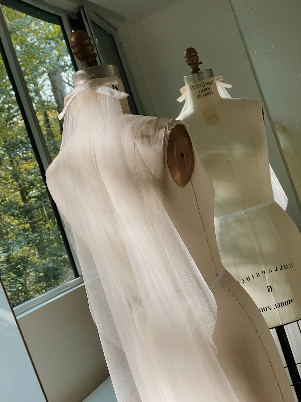 A Noémiah Bow Veil, draped over a dress form with sheer white fabric, stands by a sunlit window. Another dress form and its reflection appear in the background as sunlight highlights the delicate textures.