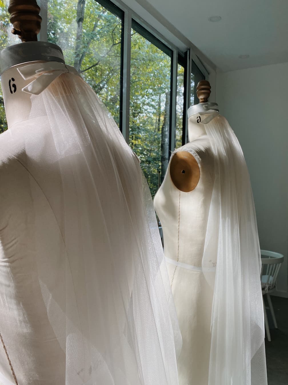 Two Noémiah Bow Veil dress forms, draped in sheer white fabric and silk bows, stand by large windows with sunlight streaming in and trees outside. The bright, modern studio showcases the elegance of the Bow Veil accessory.