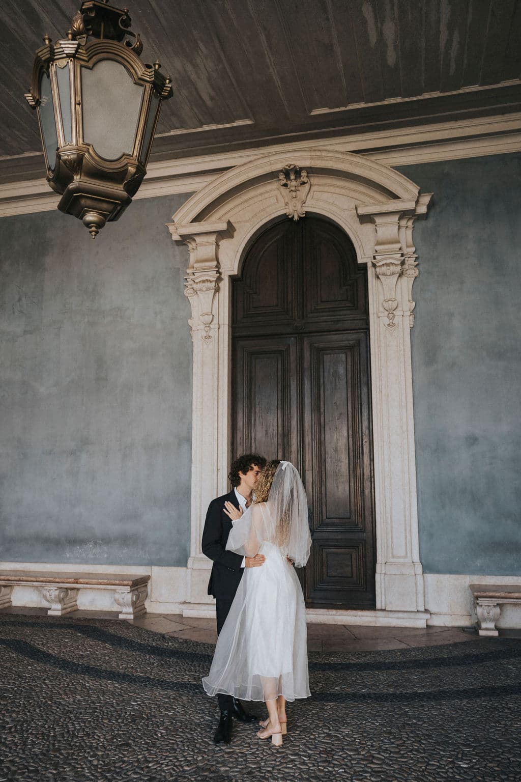 A couple embraces in front of a large, ornate wooden door. Dressed in wedding attire, the bride's Bow Veil by Noémiah flows elegantly with her white gown, while the groom stands beside her in a dark suit. A vintage hanging lantern casts a warm glow above them.