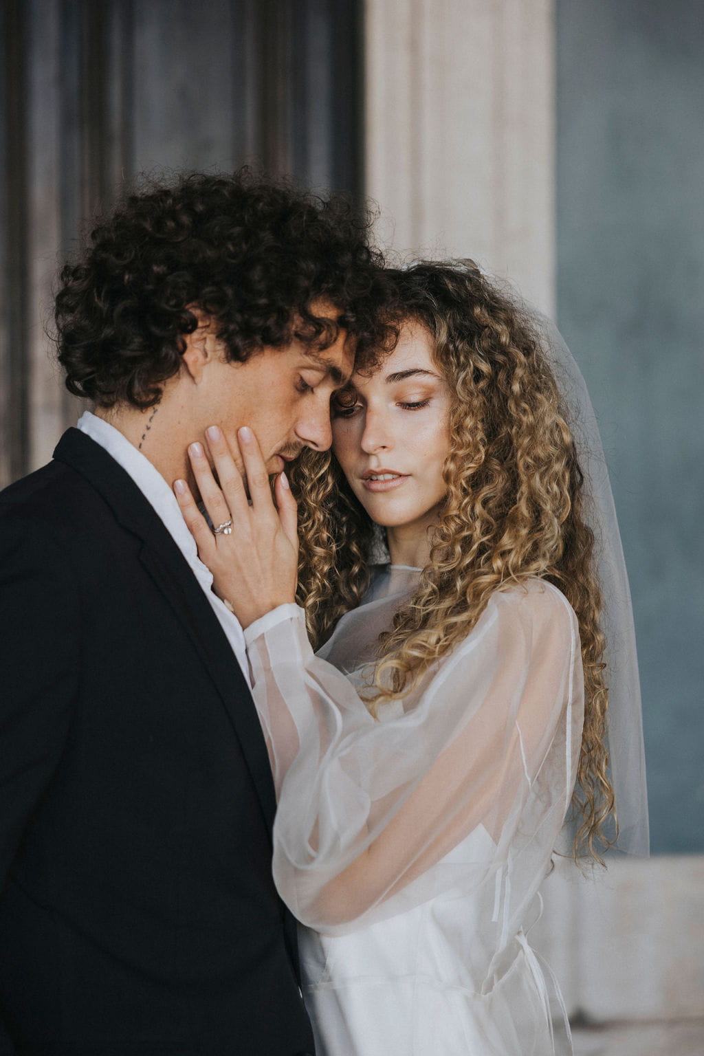 A bride and groom share an intimate moment. The bride, with long curly hair, wears a white dress complemented by the Bow Veil from Noémiah and a silk bow. The groom, sporting curly hair, is dressed in a dark suit. They gently touch foreheads as her hand rests tenderly on his face.