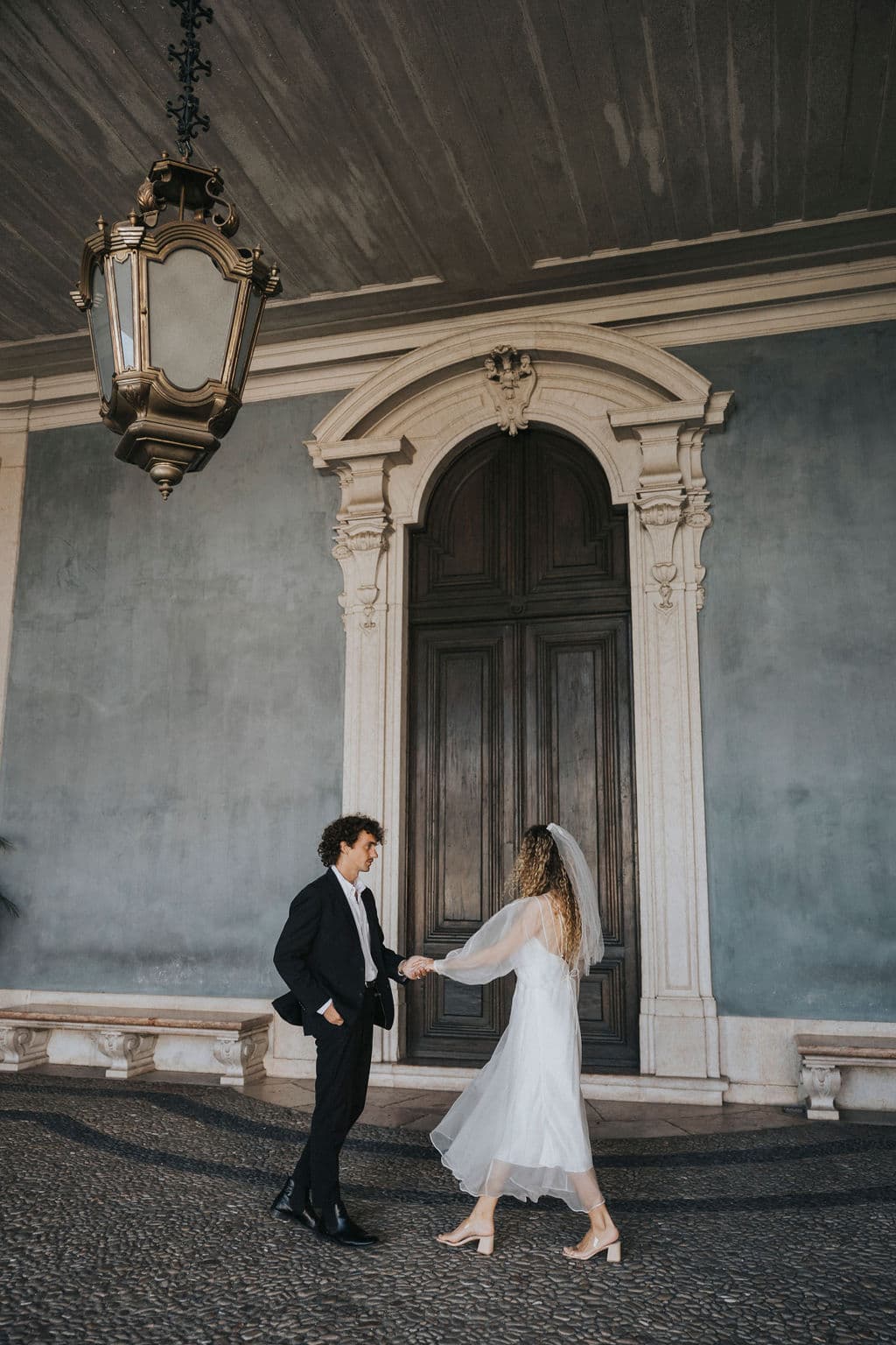 A couple holding hands stands under a large, ornate light fixture in front of an elegant wooden door. The woman is wearing Noémiah's Bow Veil and a white dress, while the man is dressed in a dark suit. The setting features a classic architectural style.