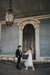 A couple holding hands stands under a large, ornate light fixture in front of an elegant wooden door. The woman is wearing Noémiah's Bow Veil and a white dress, while the man is dressed in a dark suit. The setting features a classic architectural style.