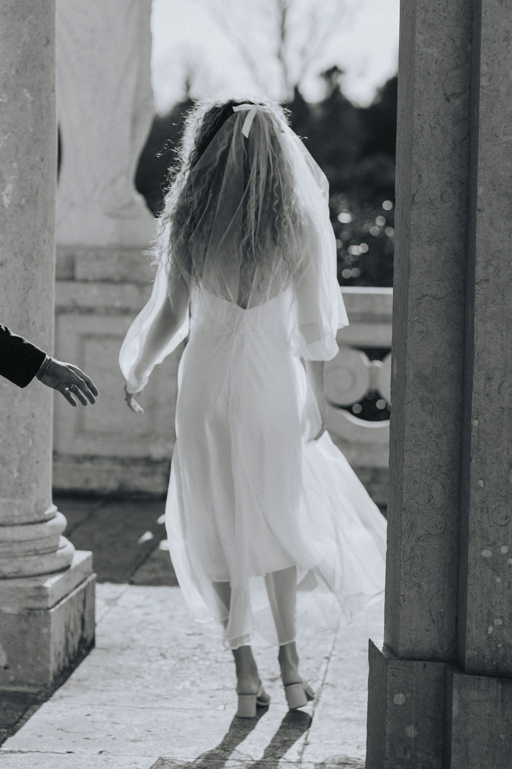 A woman in a flowing white dress, accessorized with the Bow Veil by Noémiah, walks away from the camera toward a sunlit garden, framed by columns. Her long hair cascades down her back.