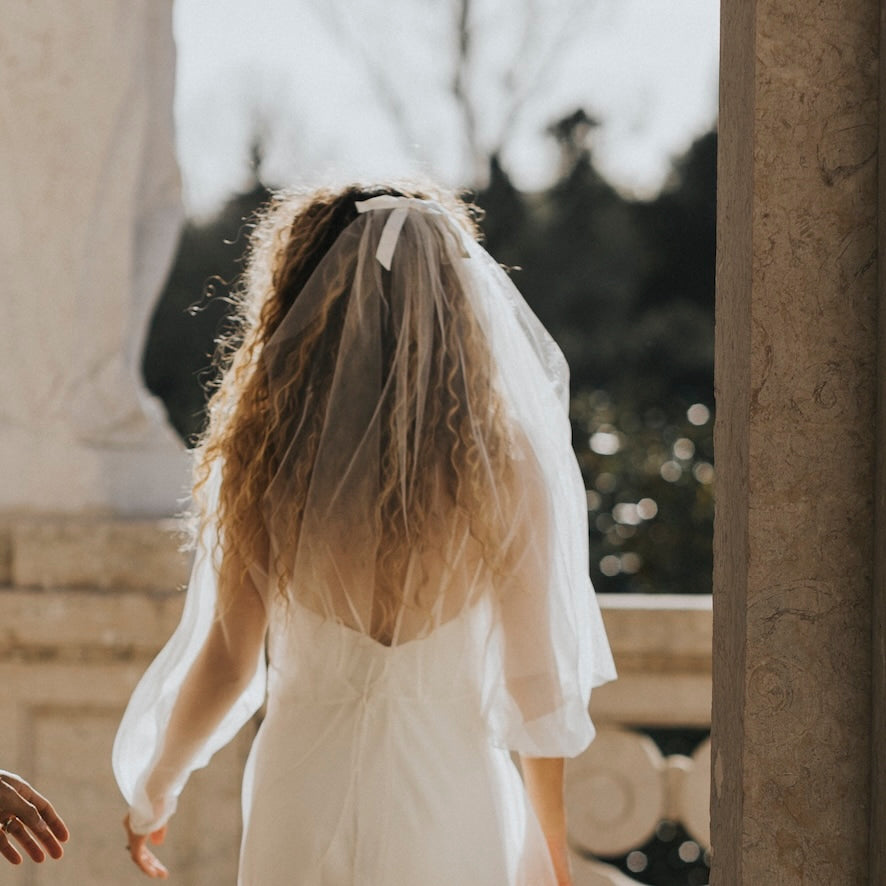 In soft sunlight, a bride with long curly hair stands outdoors, wearing a white gown adorned with Noémiah's Bow Veil. She faces away, walking towards an open space framed by stone pillars and a blurred background of trees, her veil fluttering gently behind her.