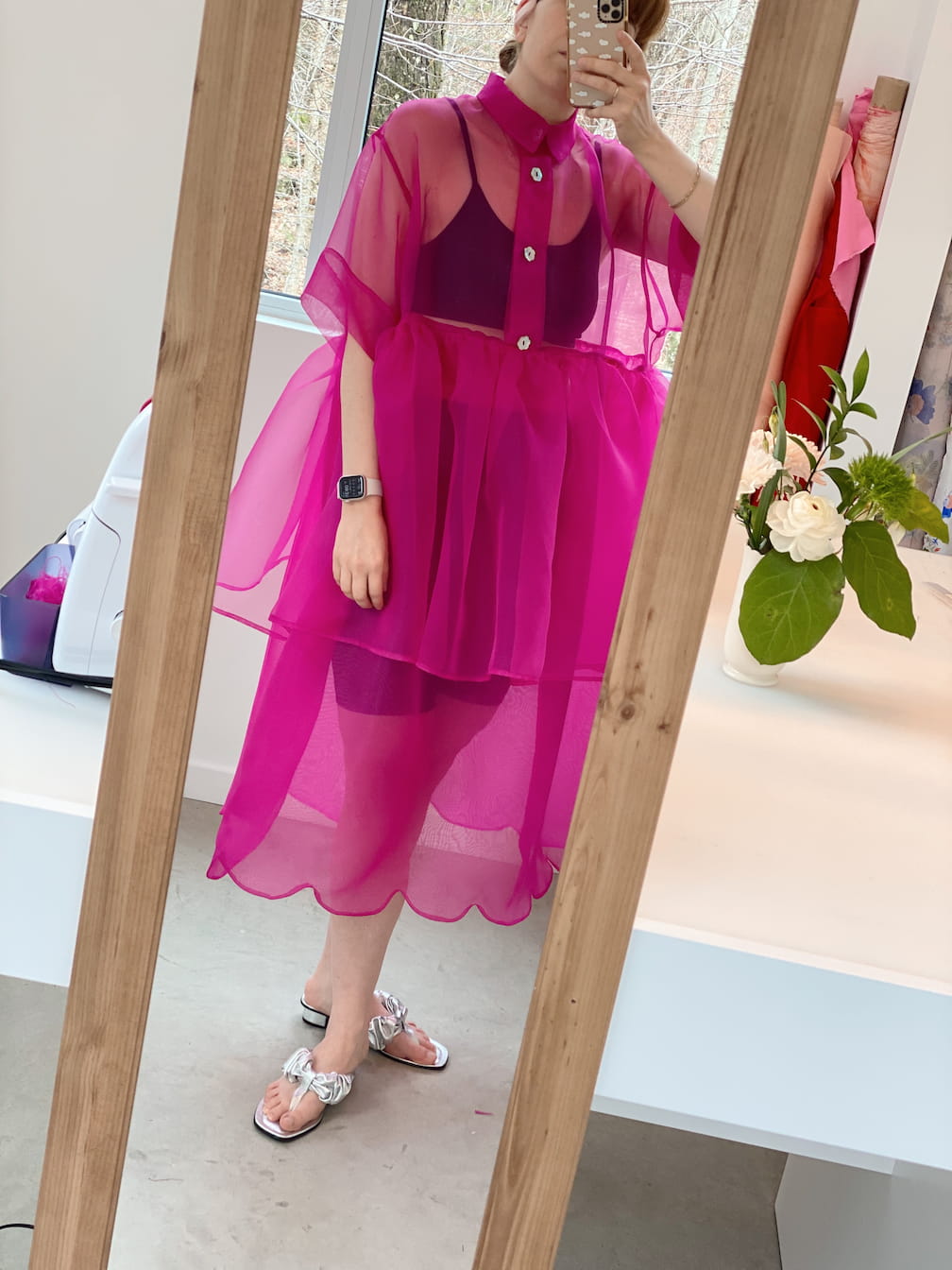 A person wearing a bright pink sheer dress and a bralette by Noémiah stands in front of a mirror, snapping a selfie. The room embraces minimalist collections with a plant on a white counter and a vacuum cleaner subtly visible in the background.