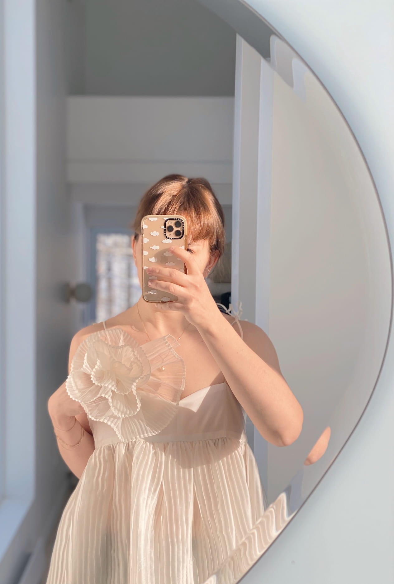 A person takes a mirror selfie with a smartphone, showcasing their light, pleated dress adorned with a hand-sewn silk organza brooch from Noémiah on the shoulder. Soft, natural light fills the space.