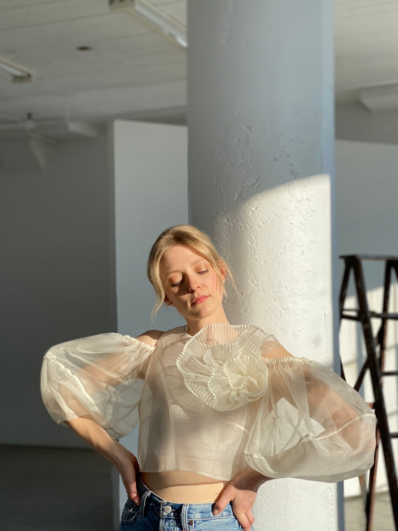 A woman stands in a sunlit room wearing Noémiah's sheer, cream-colored blouse. This elegant top is meticulously crafted from silk organza and showcases puffy sleeves with delicate floral details. With her eyes gently closed, she leans slightly against a white pillar while a wooden ladder can be seen in the background.