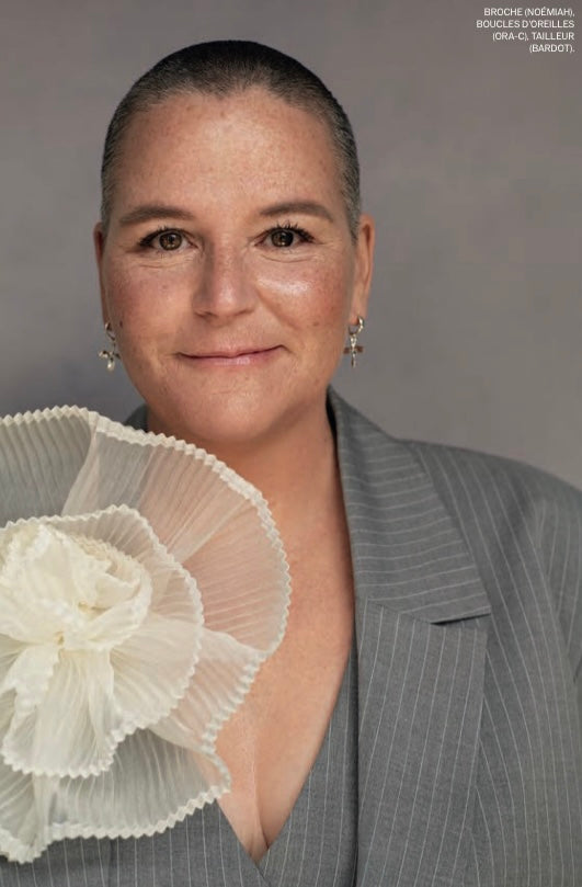 A person with short hair smiles warmly at the camera, dressed in a gray pinstripe suit jacket and holding a large white Noémiah Brooch made from silk organza. The background is a soft gray, with text visible in the upper right corner.