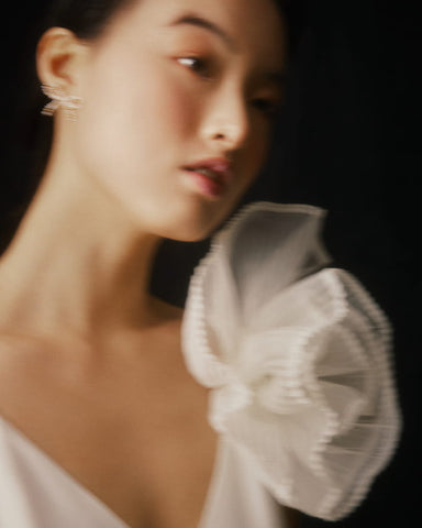 A blurred portrait showcases a person adorned in a handcrafted silk organza ensemble by Noémiah, featuring a white outfit with dramatic ruffled shoulder detail and a pleated brooch. The dreamy effect is amplified by the dark background and soft focus, while the subtle expression gains elegance from the small earring enhancing the look.