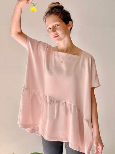 A person wearing the Camélia Top — Cap Sleeves by Noémiah, characterized by its loose and oversized boxy fit in light pink, holds a small yellow flower aloft. Their hair is tied up as they stand against a plain background, with a green plant leaf visible in the bottom corner.