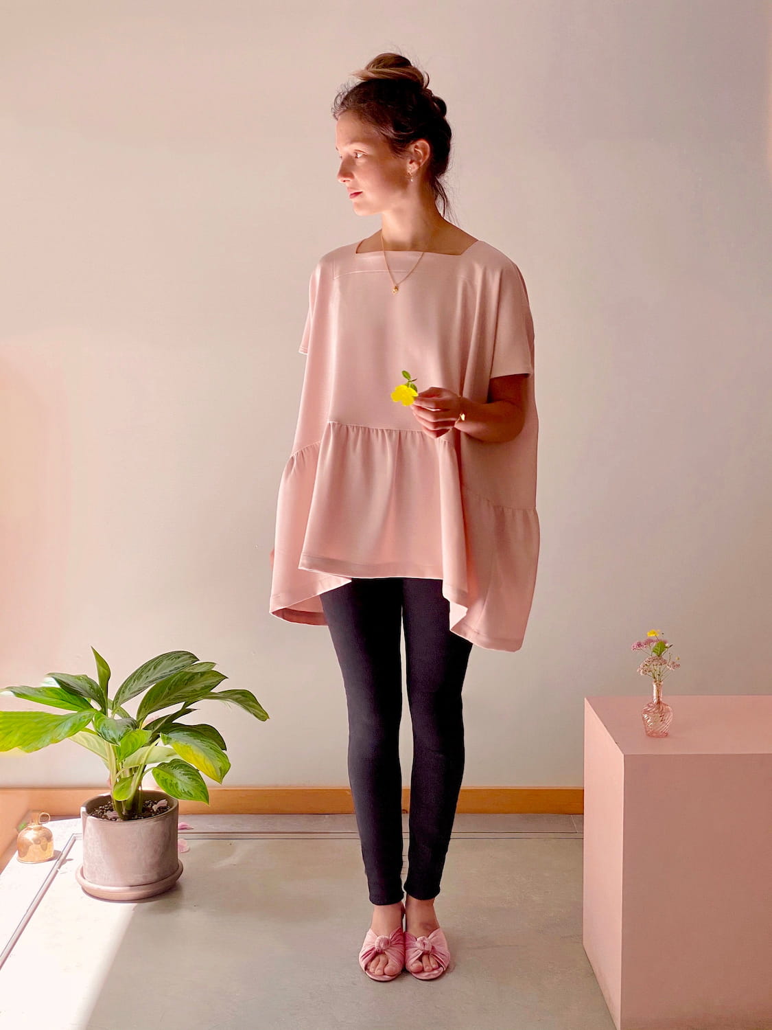 In a well-lit room, an individual holds a yellow flower while wearing the exclusive Noémiah Camélia Top with cap sleeves in pink, paired with black pants. Their hair is neatly tied up. A potted plant is positioned on the floor, and a small vase with flowers sits on a block nearby, enhancing the neutral-toned walls.