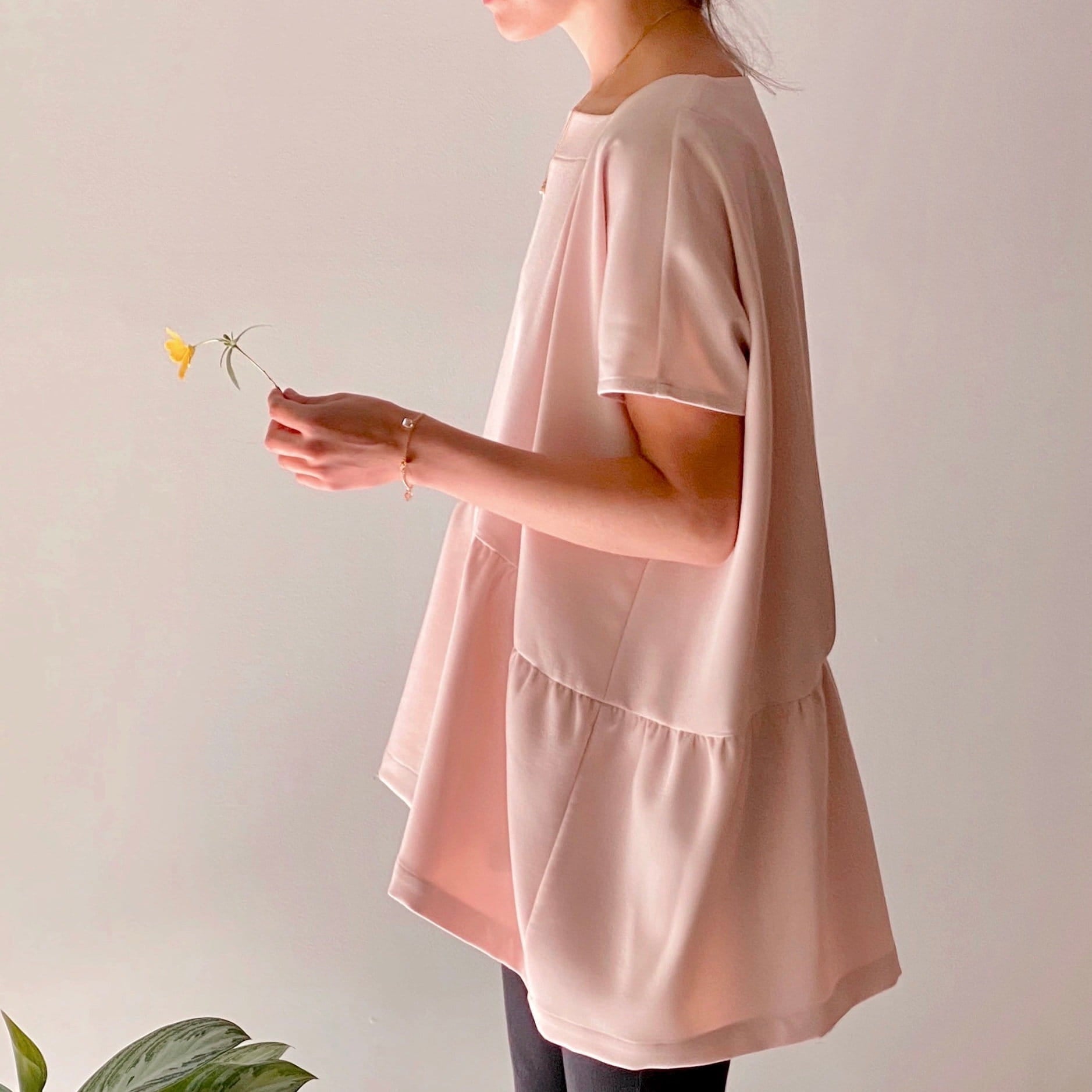 A person wearing the Camélia Top with cap sleeves by Noémiah showcases its elegant square neckline and loose-fitting design while holding a small yellow flower. The simple, soft background enhances the top's flowing elegance, and a small green plant can be seen in the bottom left corner.