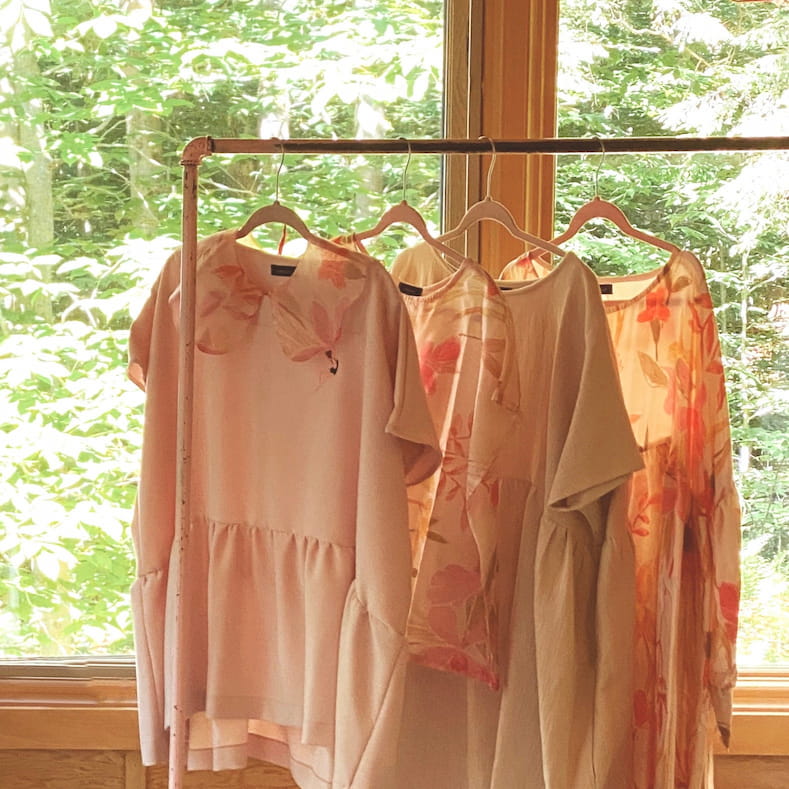 Four Camélia Tops by Noémiah, featuring cap sleeves and exclusive peach and floral embroidery, hang on a rack by a window with greenery visible outside. The light, airy fabric and subtle designs create a soft and serene atmosphere, enhancing their charm and beauty.