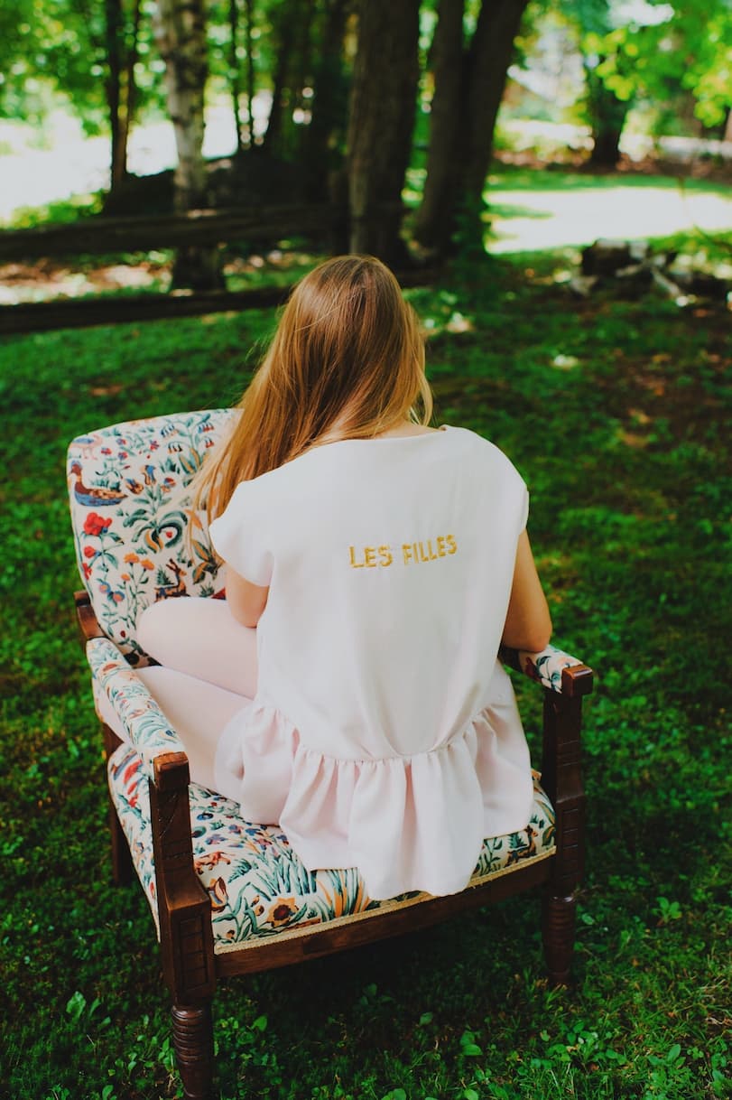 A woman with long hair sits on a floral-patterned armchair outdoors, wearing the Camélia Top — Cap Sleeves by Noémiah, featuring LES FILLES printed on the back. The light pink top's square neckline and oversized boxy shape enhance the charming scene with a grassy area and trees in the background.