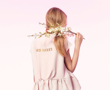 A woman with long blonde hair stands with her back to the camera, holding a branch of white flowers over her shoulder. She wears the Camélia Top — Cap Sleeves by Noémiah in a light pink hue, featuring exclusive embroidery and "LES FILLES" printed on the back. The background is a soft pink gradient.