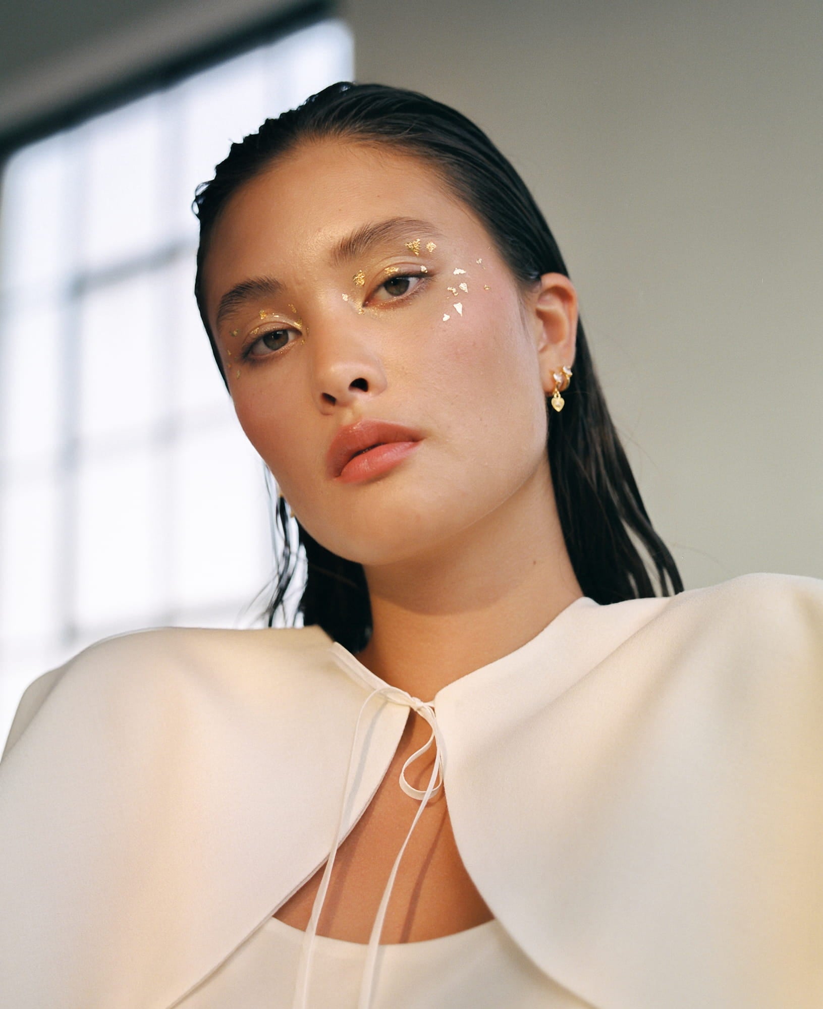 A woman with a neutral expression wears a white wool-crepe outfit, accented by the Camille Capelet from Noémiah. Her hair is slicked back, and gold decorative elements graze her eyes. She stands in front of a softly lit window, exuding the serene elegance of an autumn bride.