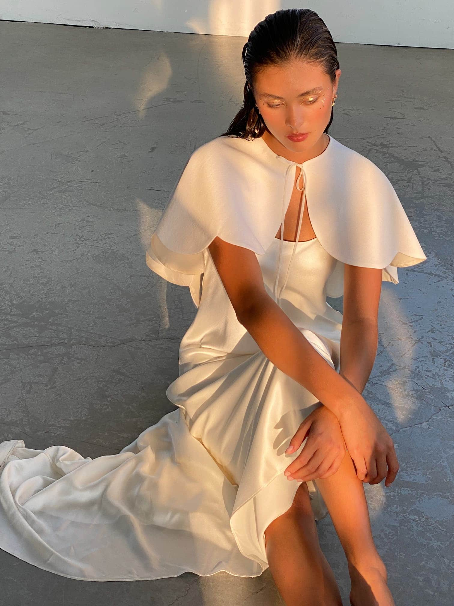 A woman sits on the floor wearing the Camille Capelet by Noémiah, a white wool-crepe dress featuring a scalloped capelet. She looks down as sunlight casts soft shadows around her in a neutral-toned room, embodying the essence of an autumn bride.