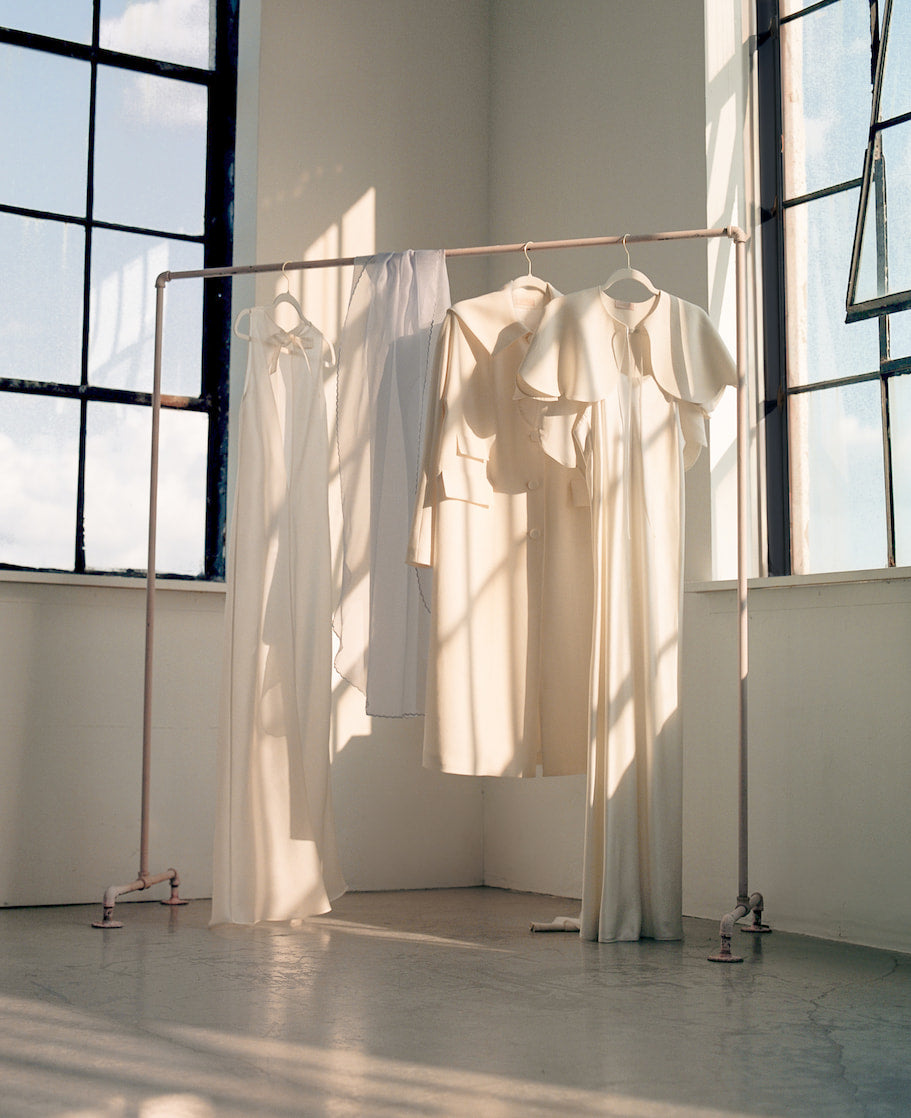 By the large windows, a clothes rack displays four Camille Capelets by Noémiah, each showcasing a long, cream-colored design with a touch of wool-crepe texture. Sunlight pours in, casting gentle shadows on the floor. Perfect for the autumn bride, these pieces embody a minimalist aesthetic with subtle elegance.