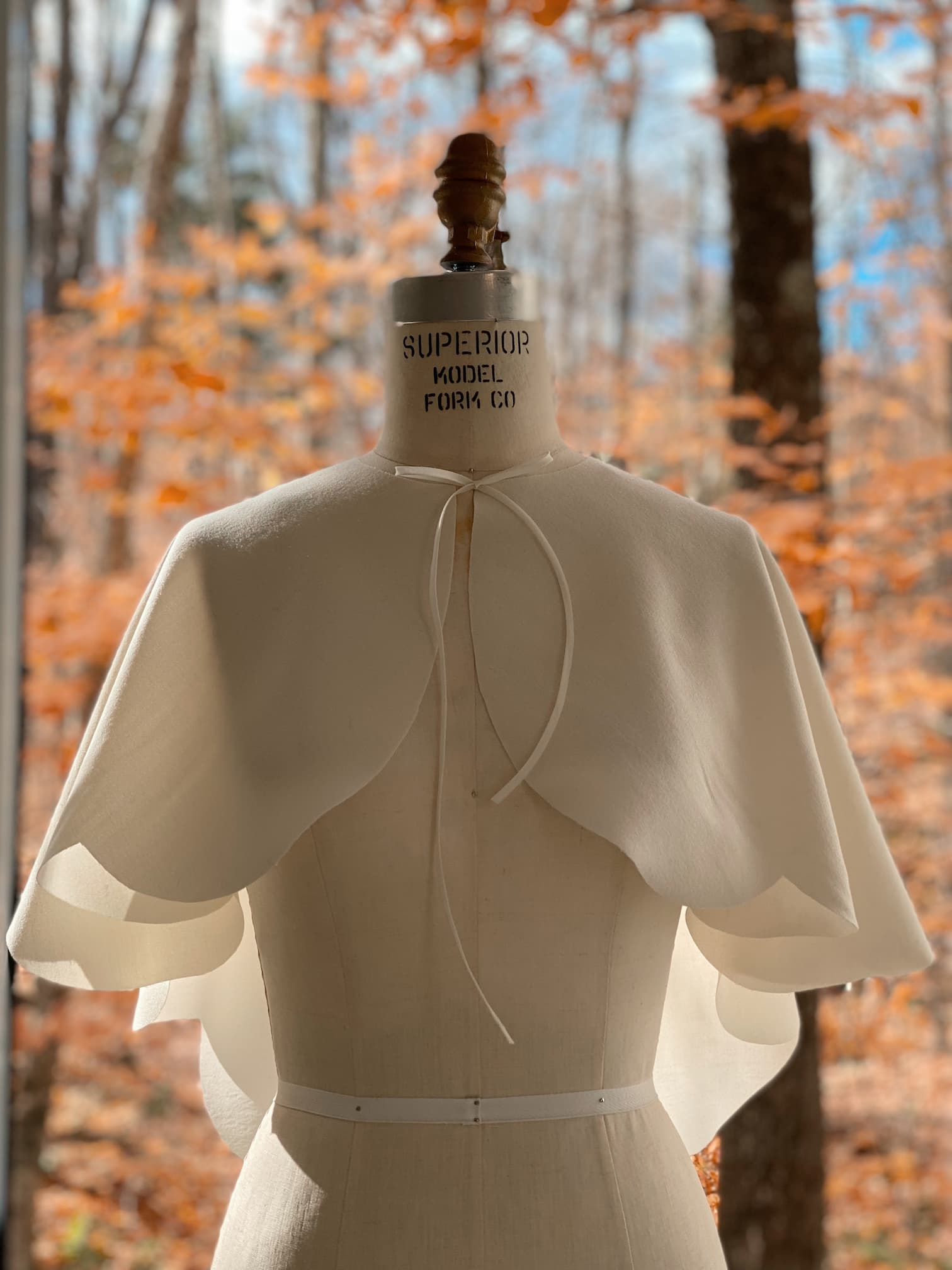 A dress form showcases a tie-back white wool-crepe fabric, elegantly complemented by the Camille Capelet from Noémiah. In the background, a forest with autumn foliage displays shades of orange and brown, creating an ideal setting for an autumn bride.