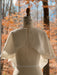 A dress form showcases a tie-back white wool-crepe fabric, elegantly complemented by the Camille Capelet from Noémiah. In the background, a forest with autumn foliage displays shades of orange and brown, creating an ideal setting for an autumn bride.