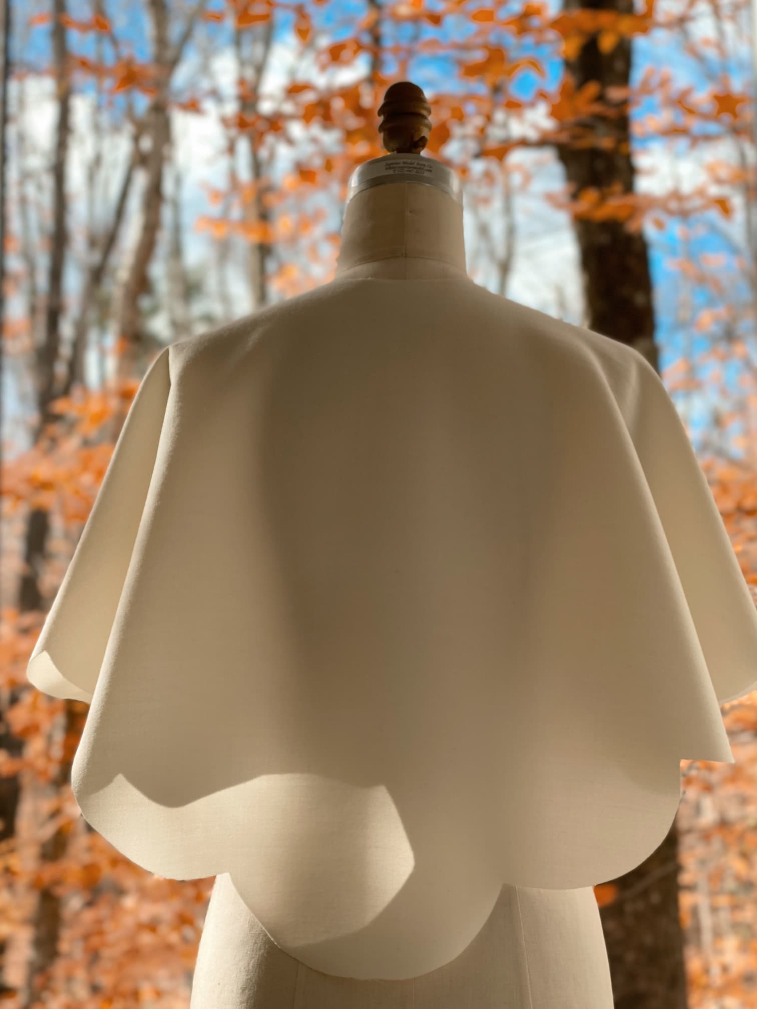 A white Camille Capelet by Noémiah is elegantly draped over a dress form, photographed outdoors. The background reveals a forest with bare trees and orange leaves, capturing the essence of an autumn bride's setting beneath a partly cloudy sky.