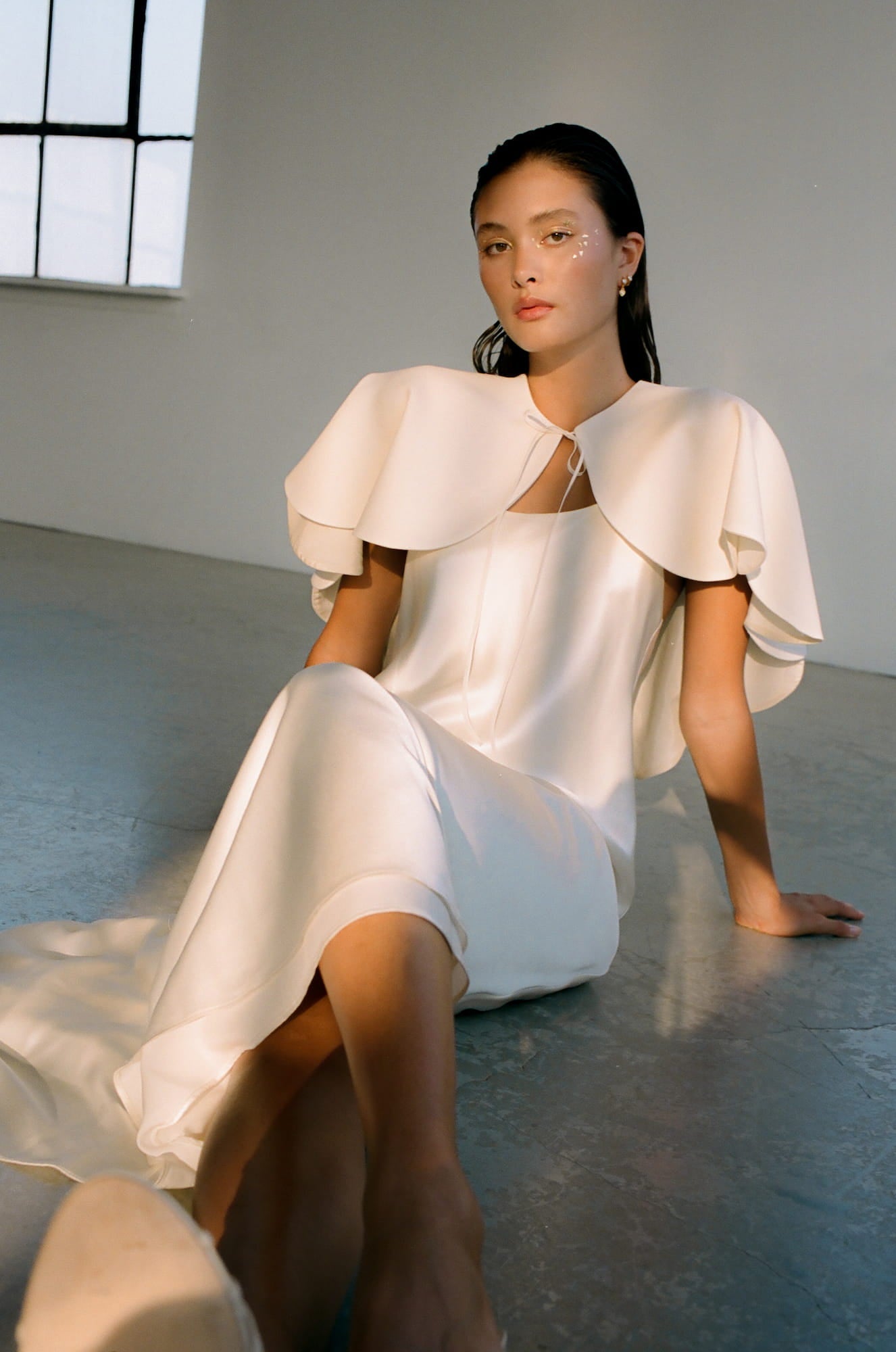 A person wearing a silky, flowing white dress with the Camille Capelet by Noémiah, featuring layered, ruffled sleeves and a scalloped design, sits on a concrete floor. Their wet-look hair and subtle face makeup complement the scene. Soft light from a large window casts an ethereal glow into the room, perfectly capturing the essence of an autumn bride.