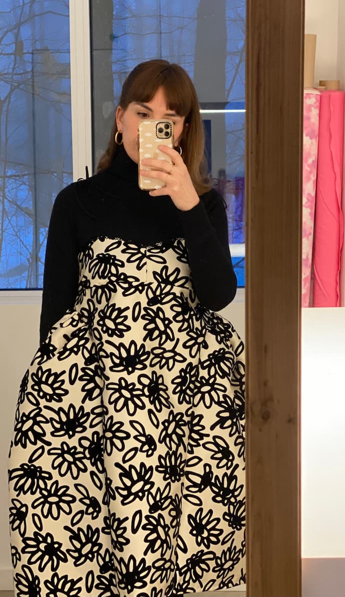 A person captures a mirror selfie wearing the Capucine Classic Dress by Noémiah, which features a bold black and white floral pattern over a black turtleneck. The snowy scenery visible through the window and shelves adorned with rolls of pink fabric lend an extra layer of depth to this chic winter ensemble.