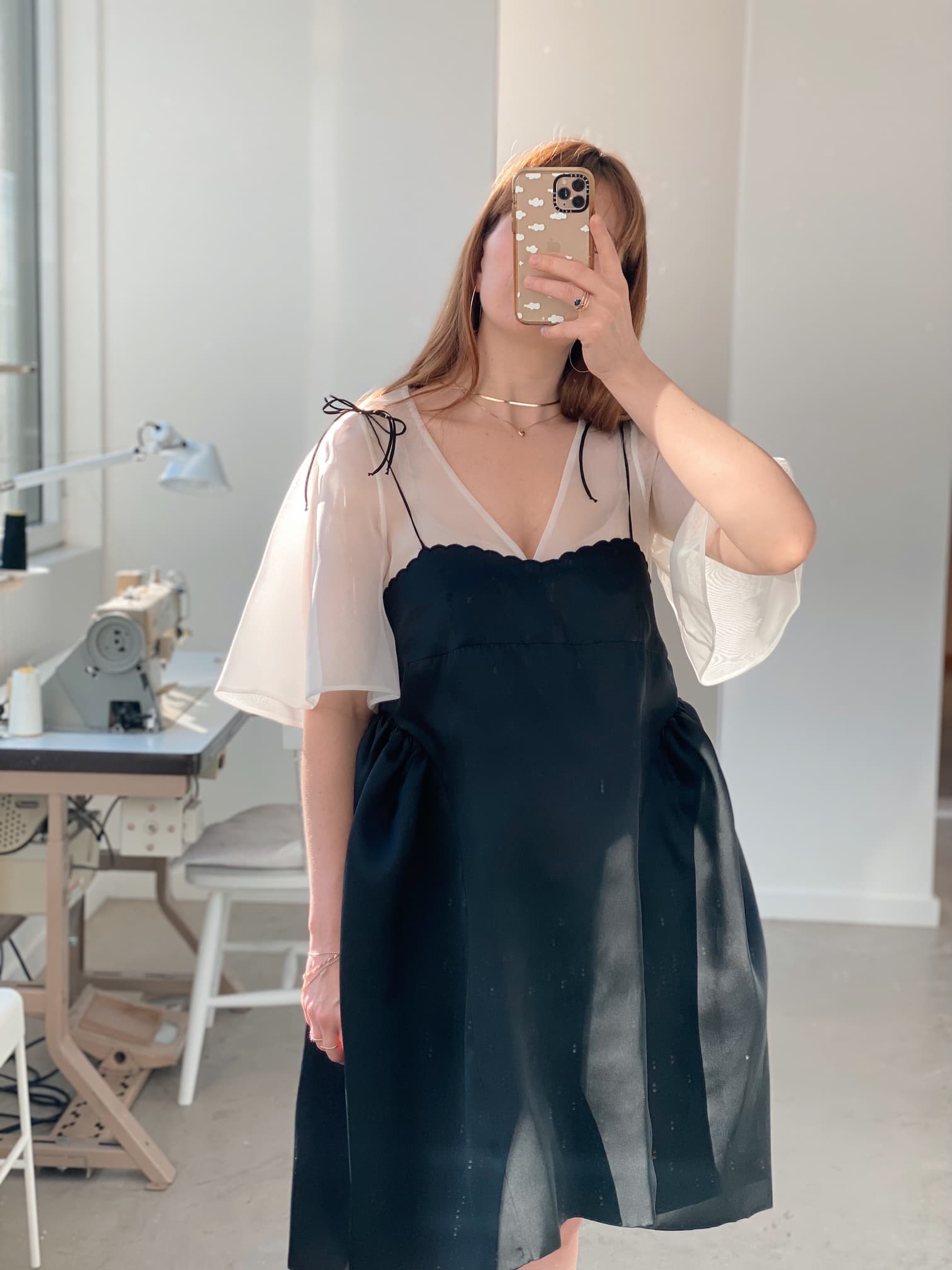 A woman takes a mirror selfie while wearing the Capucine Classic Dress from Noémiah, featuring sheer white sleeves that evoke the look of a silk mini dress. The sewing machine in the background suggests the intricate craftsmanship, such as scalloped embroidery, that crafted this elegant ensemble.