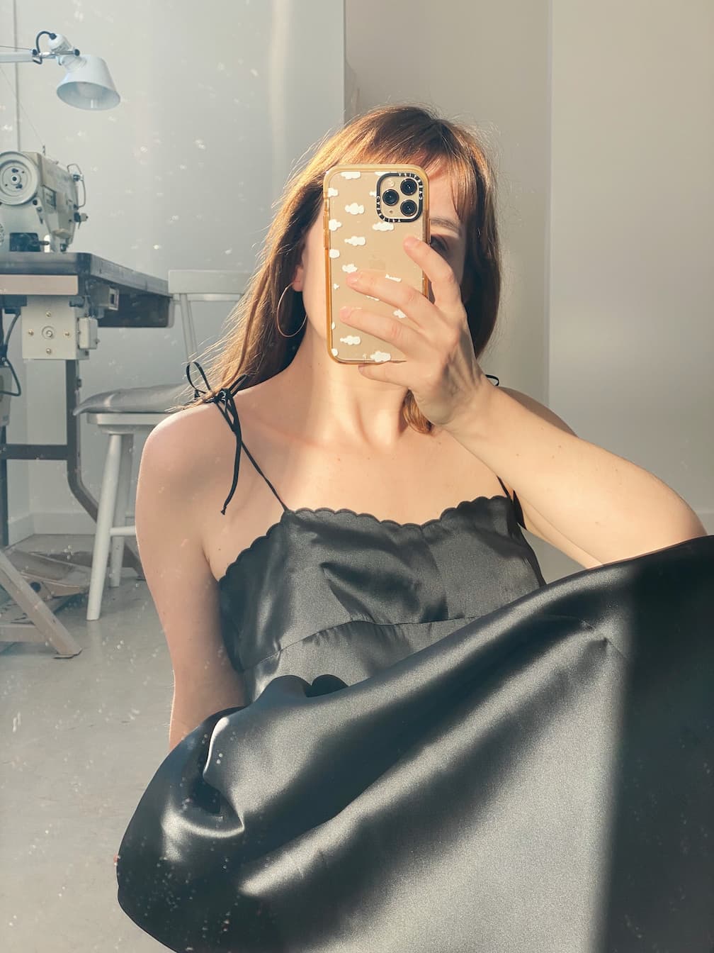 A person poses in a mirror selfie, highlighting the Capucine Classic Dress by Noémiah, a black satin bustier mini dress adorned with delicate scalloped embroidery. The well-lit room reveals a sewing machine and stool, while the phone case features a cloud pattern.