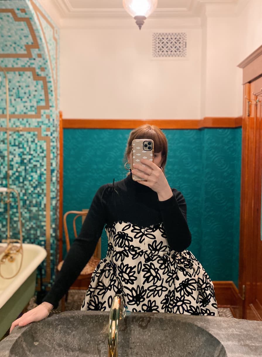 A person wearing a black top and the Capucine Classic Dress by Noémiah, which is a black and white patterned bustier mini dress with silk shoulder straps, takes a mirror selfie in a bathroom. The space features teal tiled walls and a vintage bathtub under a decorative ceiling light.