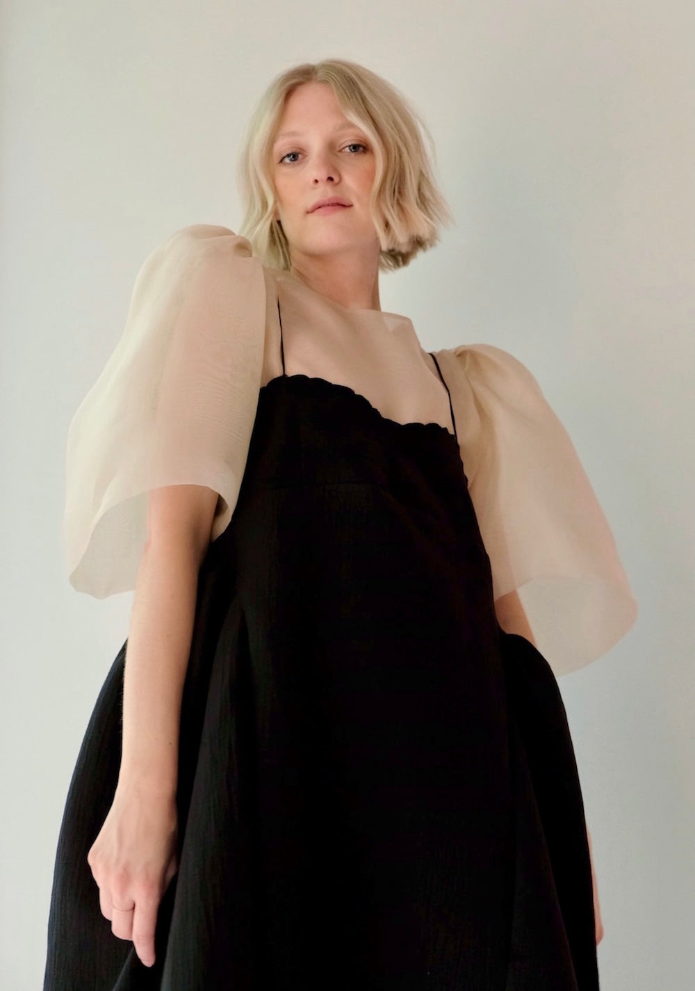 A person with short blond hair is wearing the Capucine Classic Dress by Noémiah, which is a bustier mini dress featuring sheer, puffy sleeves and a layered black skirt. The dress is accentuated with scalloped embroidery on its silk shoulder straps. They are standing against a plain, light background, gazing directly at the camera.