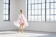 A woman in a Capucine Floral Dress by Noémiah, paired with a white shawl, stands barefoot in a bright room with large windows. Sunlight streams in, casting shadows on the floor. A bridge is partially visible through the windows, highlighting the elegance of her custom-made attire.