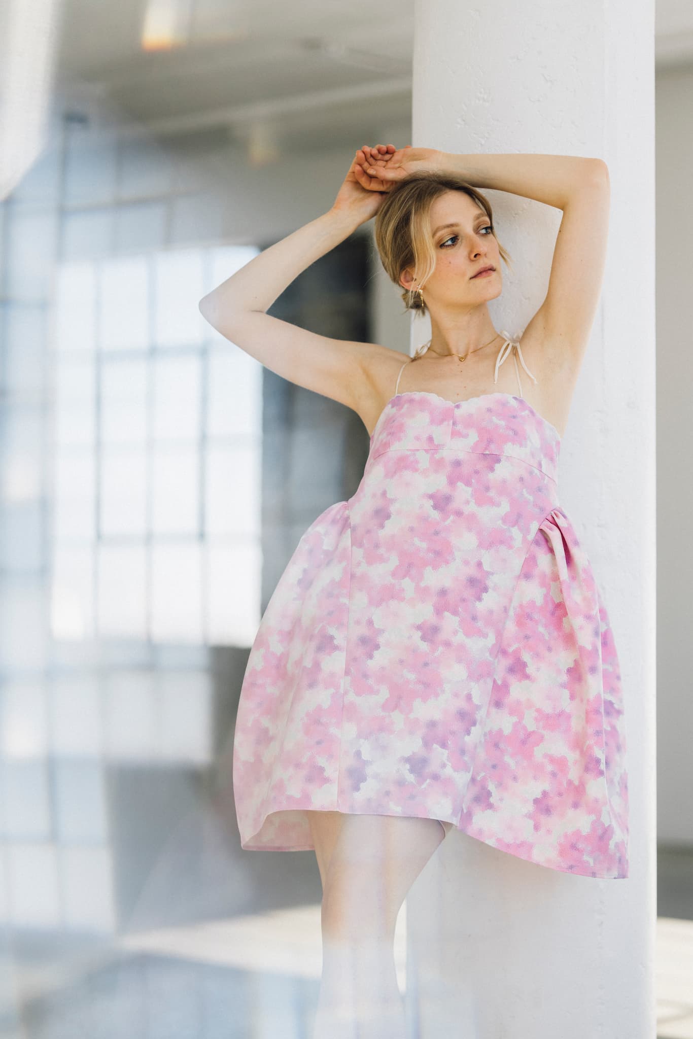 A woman in a Noémiah Capucine Floral Dress, featuring a pink and white floral bustier design, leans against a white pillar. The deadstock silk fabric with thin straps and a full skirt enhances her upward gaze. An interior window softly frames the airy atmosphere in the background.