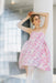 A woman in a Noémiah Capucine Floral Dress, featuring a pink and white floral bustier design, leans against a white pillar. The deadstock silk fabric with thin straps and a full skirt enhances her upward gaze. An interior window softly frames the airy atmosphere in the background.