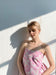 A person wearing the Capucine Floral Dress by Noémiah, a pink and white bustier mini dress, stands against a sunlit wall. Their eyes are closed, and soft shadows create patterns around them in the delicate folds of deadstock silk.