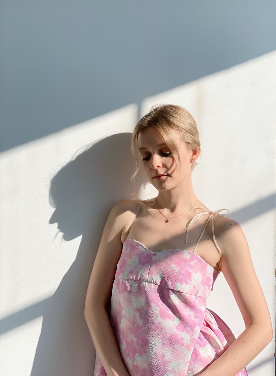 A person wearing the Capucine Floral Dress by Noémiah, a pink and white bustier mini dress, stands against a sunlit wall. Their eyes are closed, and soft shadows create patterns around them in the delicate folds of deadstock silk.