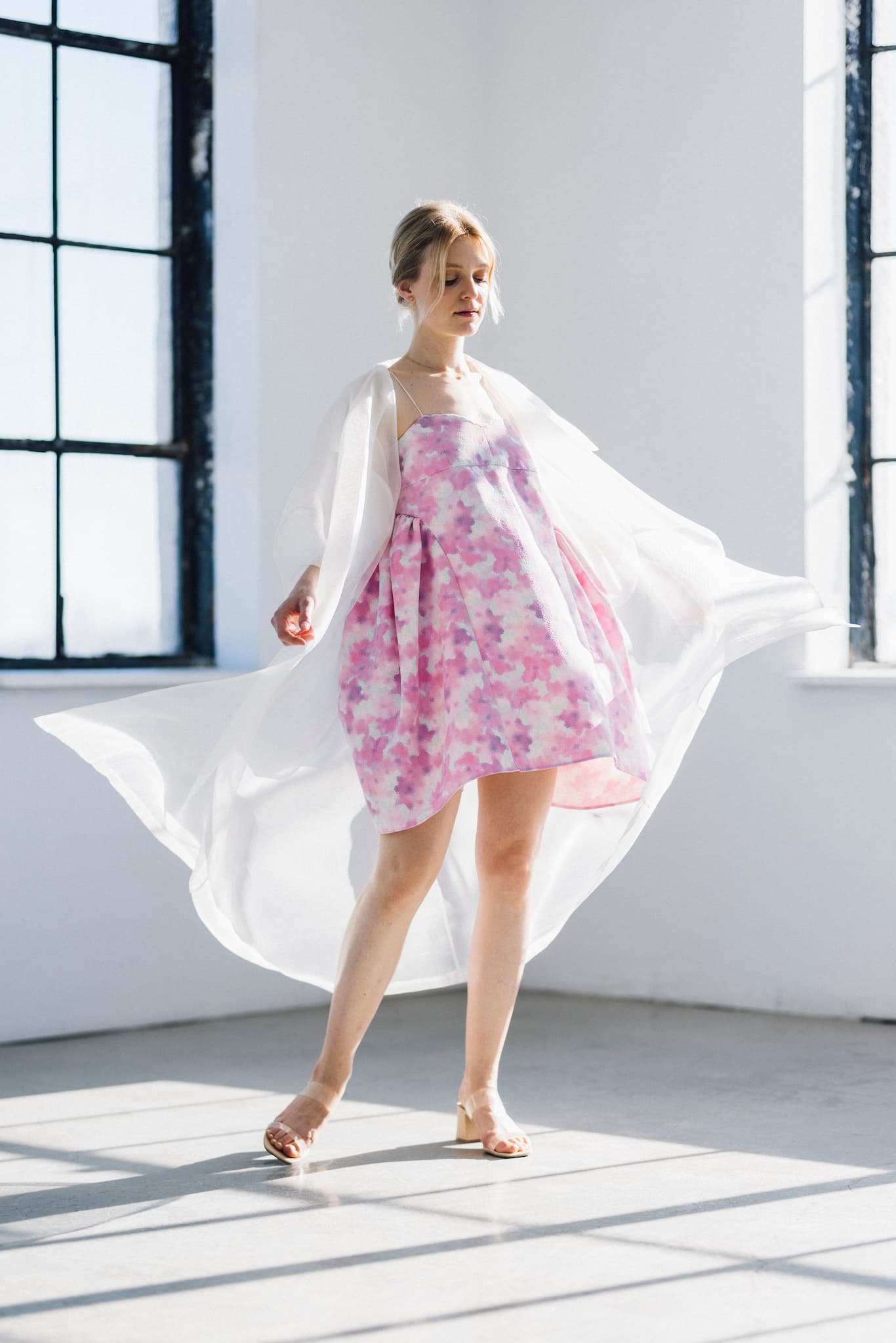 In a bright room with large windows, a woman is wearing the Capucine Floral Dress by Noémiah, featuring pink and purple florals on deadstock silk, elegantly complemented by a sheer white overlay. Her light-colored hair is styled up, and she completes the ensemble with sandal heels as sunlight casts soft shadows on the floor.