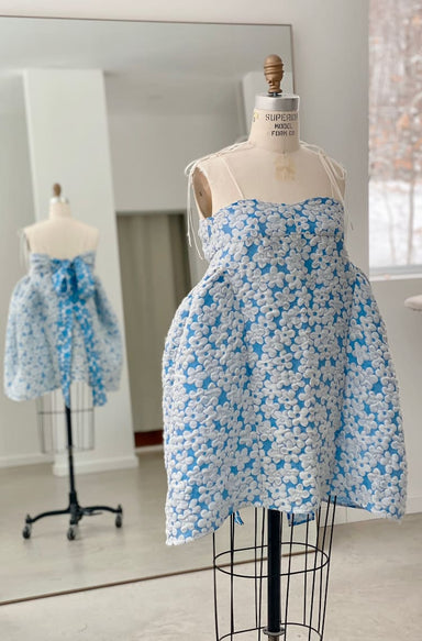 In a bright and minimalistic room, a dress form elegantly displays the Capucine Floral Dress by Noémiah, crafted from blue and white floral deadstock silk. The dress features a voluminous skirt, tied straps, and a bow at the back, all beautifully reflected in the mirror. This made-to-measure piece exudes sophistication.