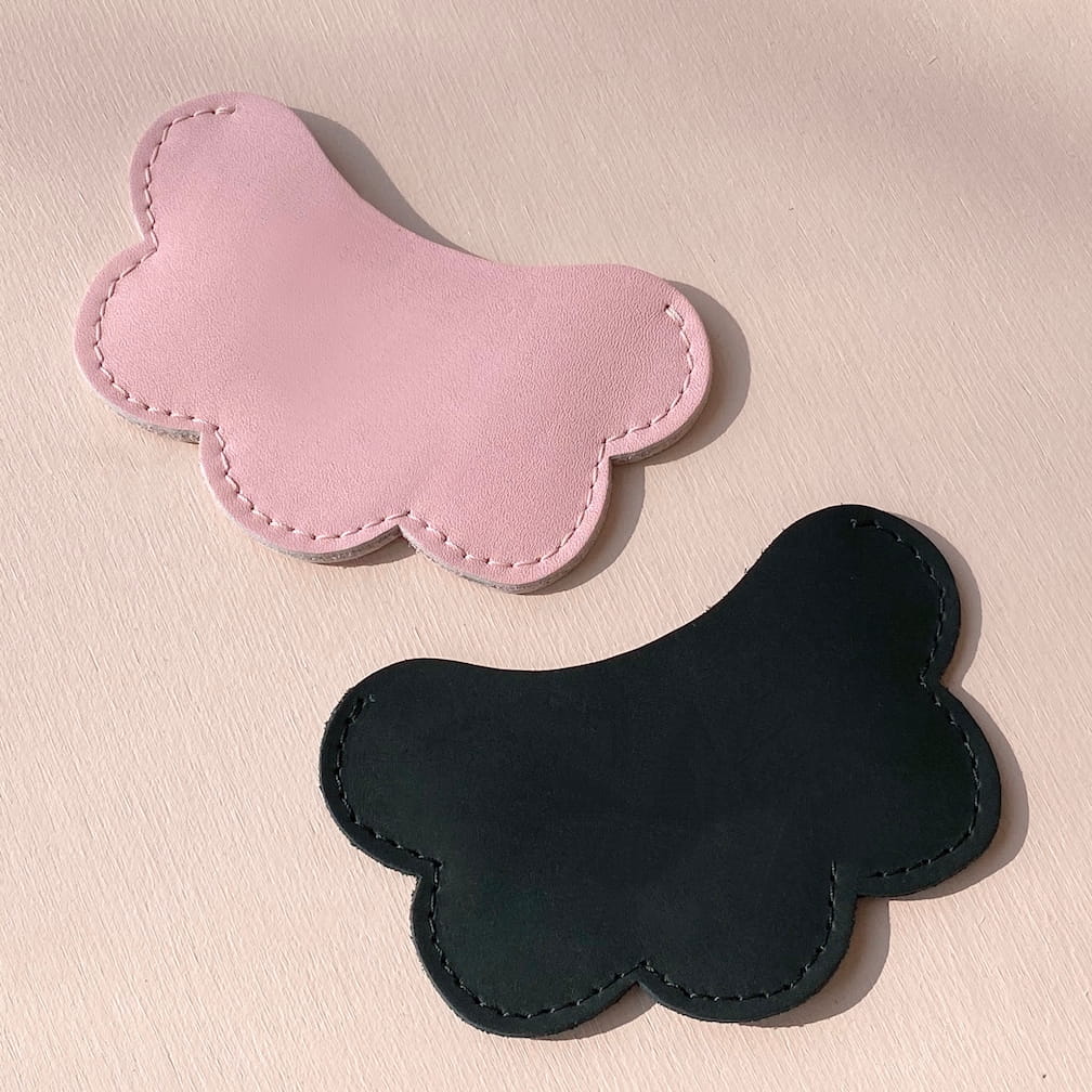 On a light beige surface, two Noémiah cloud-shaped card holders, one in pink and the other in black leather, feature a distinctive scalloped design with visible stitching along the edges.