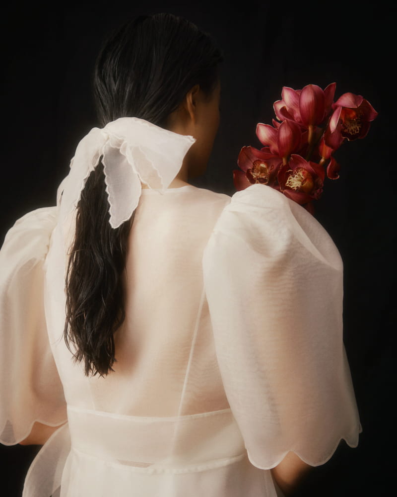 A woman with long dark hair, tied with a sheer white ribbon, faces away. She wears the Cassandre Wrap Dress by Noémiah, a light puffy-sleeved wrap dress made of silk organza, and holds a bouquet of vibrant red orchids over her left shoulder against a dark background.