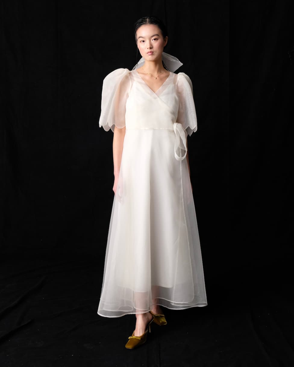 A woman stands against a black background in a handmade-to-order Cassandre Wrap Dress by Noémiah, featuring a long sheer white design with puffed sleeves and a ribbon at the waist. She pairs the silk organza outfit with green velvet shoes, her hair tied back neatly.