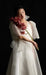 Wearing the Cassandre Wrap Dress by Noémiah, a person stands out against a black background, holding a stem of deep red orchids. The flowing white gown's translucent silk organza sleeves accentuate its handmade-to-order craftsmanship, while the gentle lighting imbues the scene with an ethereal quality.