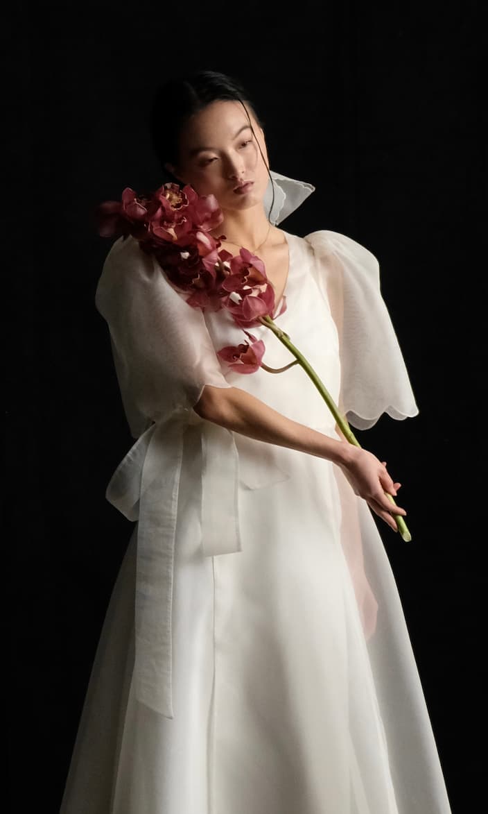 Wearing the Cassandre Wrap Dress by Noémiah, a person stands out against a black background, holding a stem of deep red orchids. The flowing white gown's translucent silk organza sleeves accentuate its handmade-to-order craftsmanship, while the gentle lighting imbues the scene with an ethereal quality.