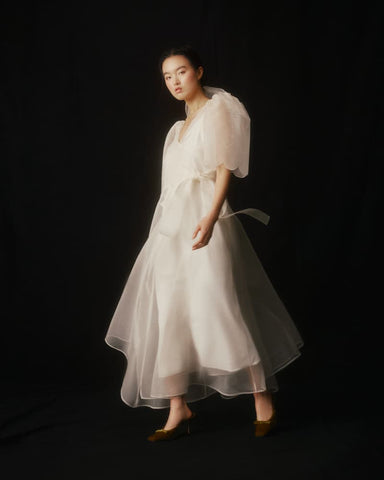 A woman stands against a black background, adorned in the Cassandre Wrap Dress by Noémiah, crafted from bespoke silk organza with puffy sleeves and a ribbon. Her shoulder-length hair enhances her elegant pose, and she completes the look with brown shoes, exuding poise and grace.