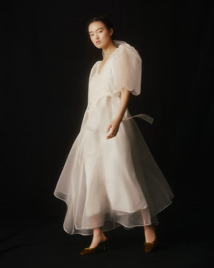 A woman stands against a black background, adorned in the Cassandre Wrap Dress by Noémiah, crafted from bespoke silk organza with puffy sleeves and a ribbon. Her shoulder-length hair enhances her elegant pose, and she completes the look with brown shoes, exuding poise and grace.