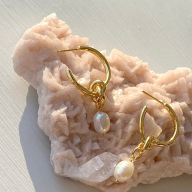 The Catherine Earrings by Noémiah, adorned with dangling freshwater pearls and gold plating, are showcased on a textured, light pink stone surface. Featuring stainless steel posts, these earrings are illuminated by natural light, accentuating their shiny appearance.