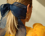 A woman with blonde hair tied in a ponytail featuring a large, sheer blue bow wears Noémiah's Catherine Earrings, gold-plated hoops adorned with freshwater pearls. She complements this elegant look with a sheer, ruffled yellow blouse, capturing attention from the back of her head and shoulder.