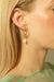 Close-up of a woman adorned with the Catherine Earrings by Noémiah, showcasing elegant gold-plated hoops with freshwater pearls. Her blonde hair is tucked behind her ear, enhancing her light, translucent green top against a neutral background.